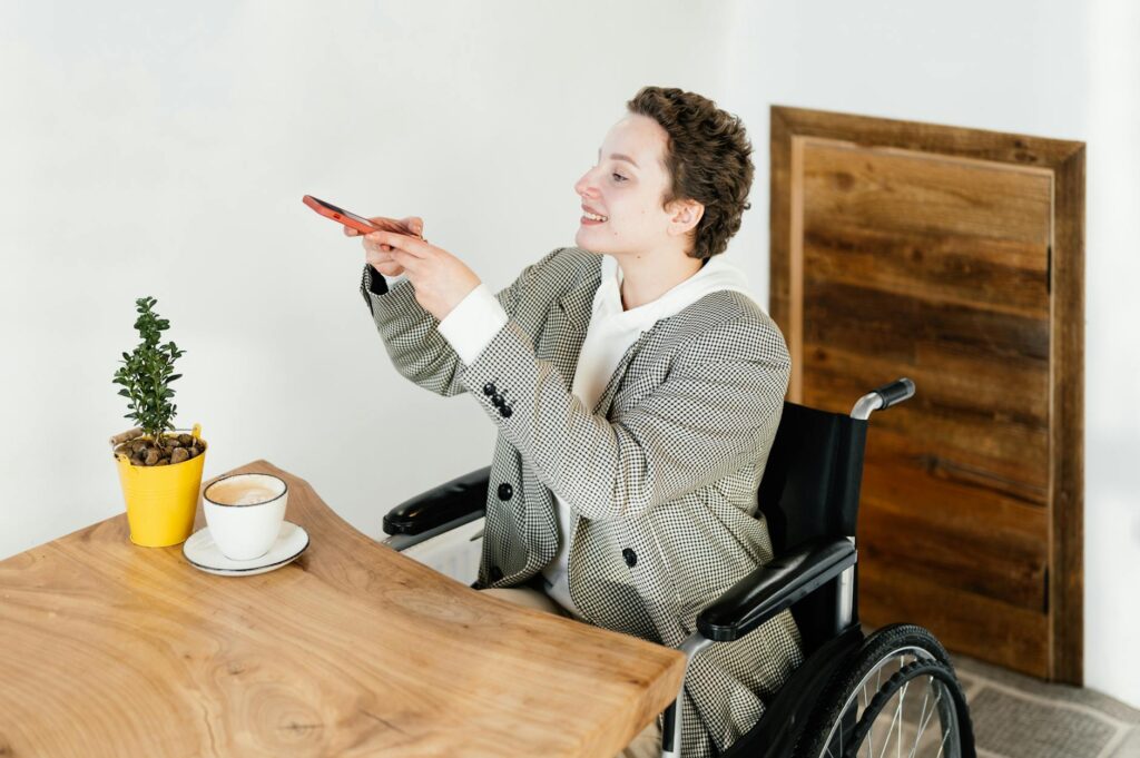 Positive female in wheelchair taking picture of hot coffee on smartphone while sitting at table with plant in modern cafe