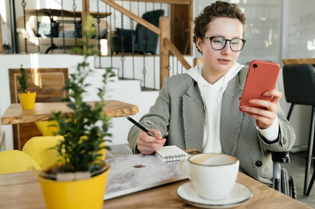 Concentrated businesswoman in formal wear and eyeglasses using wheelchair browsing smartphone and taking notes in copybook while working in cafe