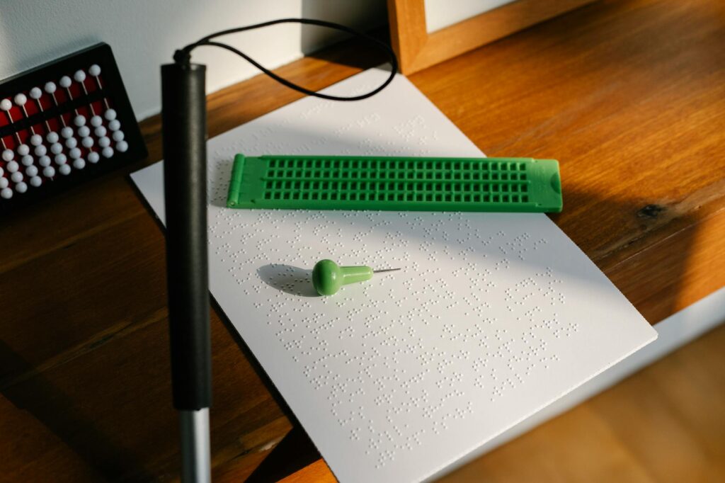 Braille sheet with stylus and cane on wooden desk in sunlight.