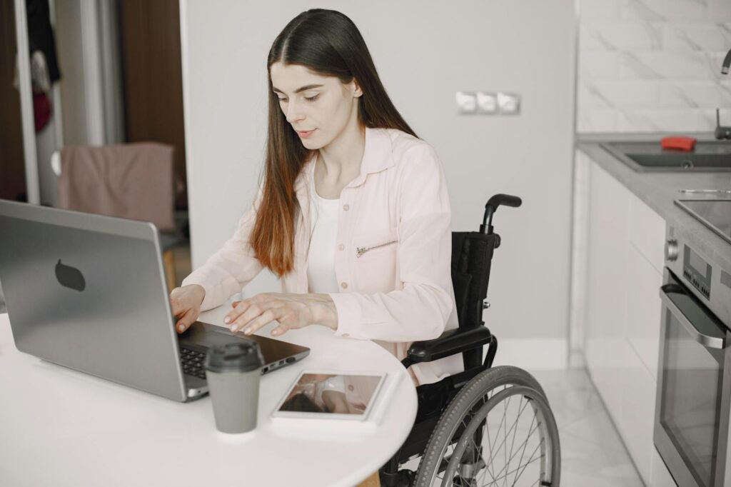 Woman in wheelchair working on laptop at home with coffee and tablet on table.