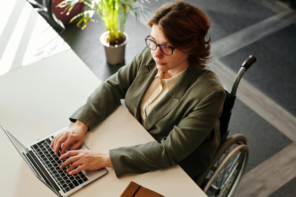 Professional woman in wheelchair working on laptop at a desk in an office setting, promoting inclusivity.