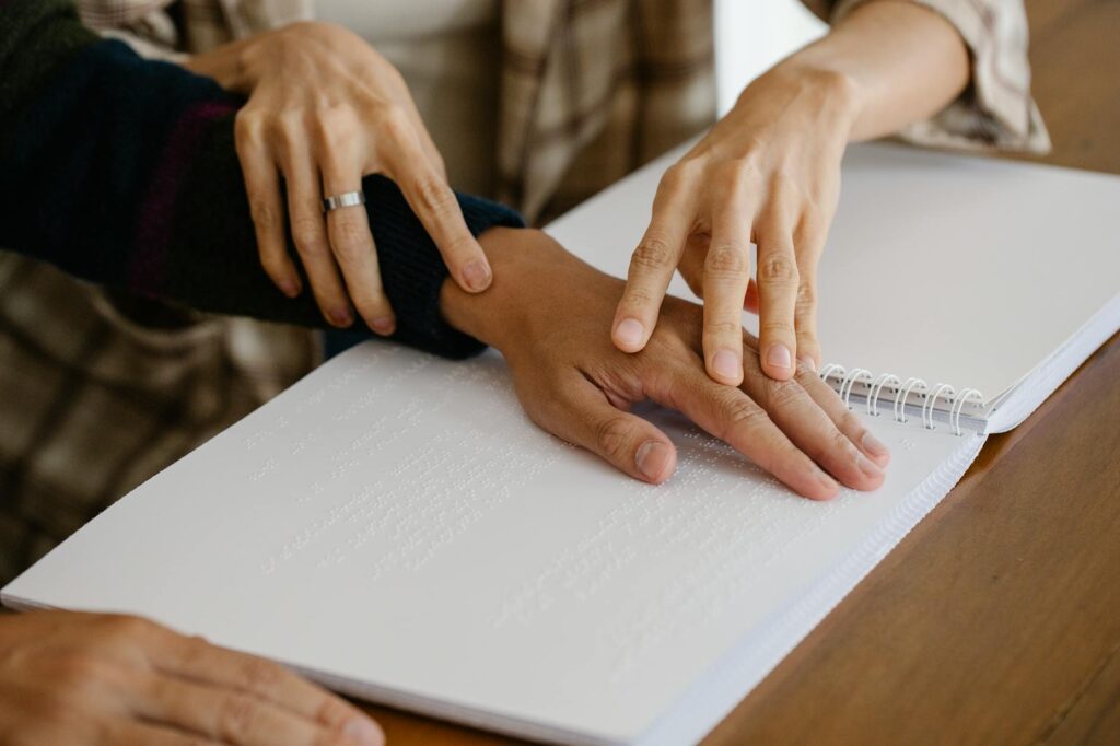Crop anonymous caregiver guiding hand of blind patient reading information in copybook with braille text